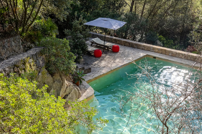 Vue surélevée d'une piscine turquoise avec terrasse en pierre, chaises longues et poufs rouges dans une forêt.