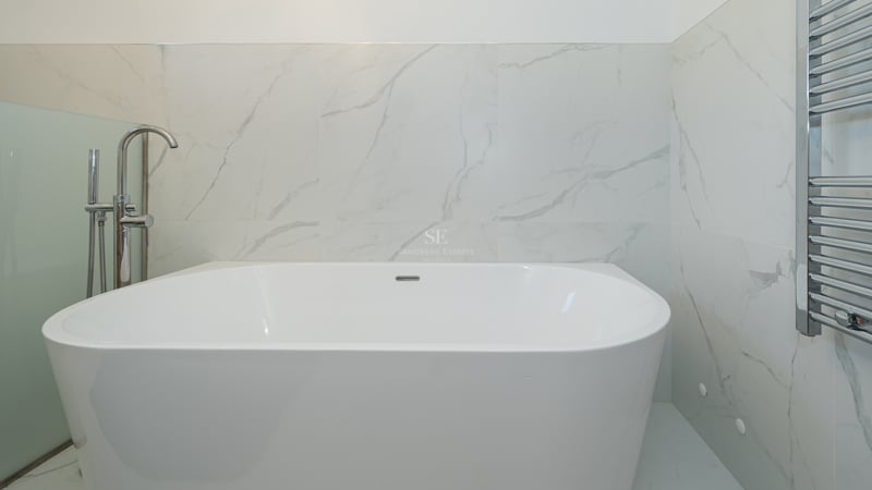 Baignoire îlot blanche avec robinetterie chromée sur fond de murs en marbre et sèche-serviettes.