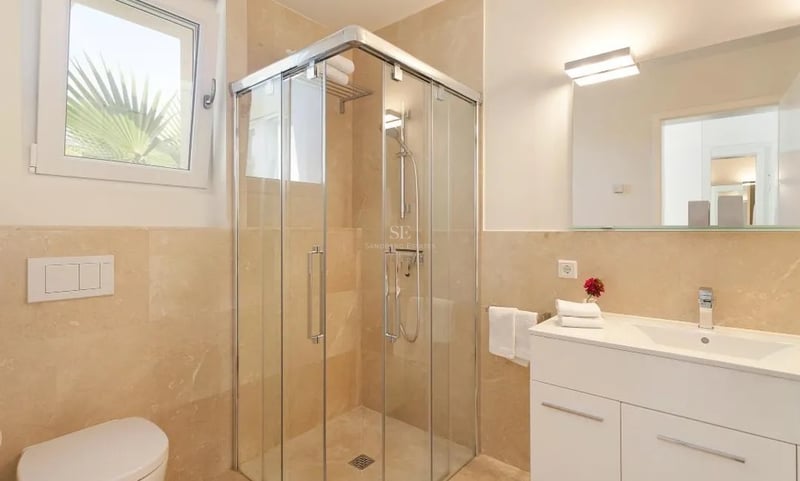 Salle de bain lumineuse avec douche d'angle en verre, carrelage en marbre beige et meuble vasque blanc.