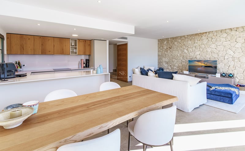 Pièce lumineuse à aire ouverte avec table à manger en bois, cuisine moderne, canapé blanc et mur en pierre naturelle.