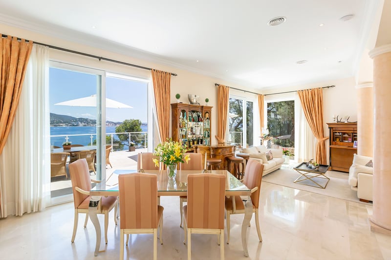Salle à manger à aire ouverte avec table en verre, sol en marbre et grandes baies vitrées donnant sur la mer.
