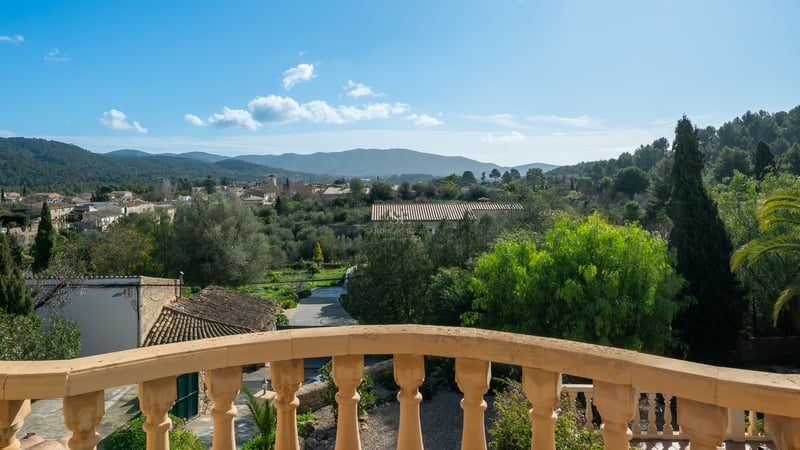 Vue depuis un balcon en pierre sur une vallée verdoyante, un village méditerranéen et des montagnes lointaines.