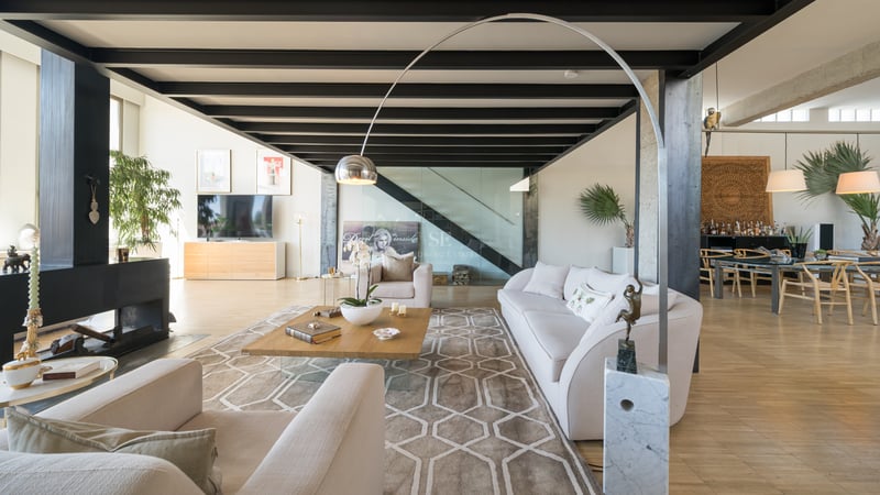 Salon moderne spacieux avec poutres en acier noir, canapé crème, tapis géométrique et escalier vitré.