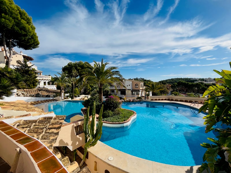 Grande piscine de style lagon avec îlot central, palmiers et villas aux toits de terre cuite sous un ciel dégagé.