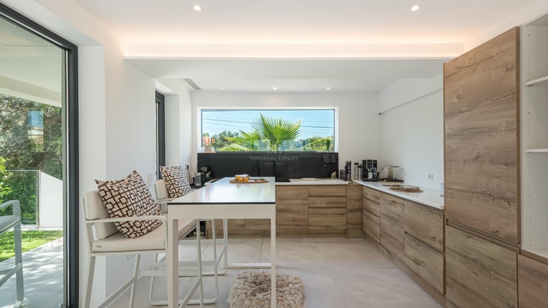 Cuisine contemporaine lumineuse avec des meubles en bois clair, un îlot blanc et une fenêtre cadrant des palmiers.