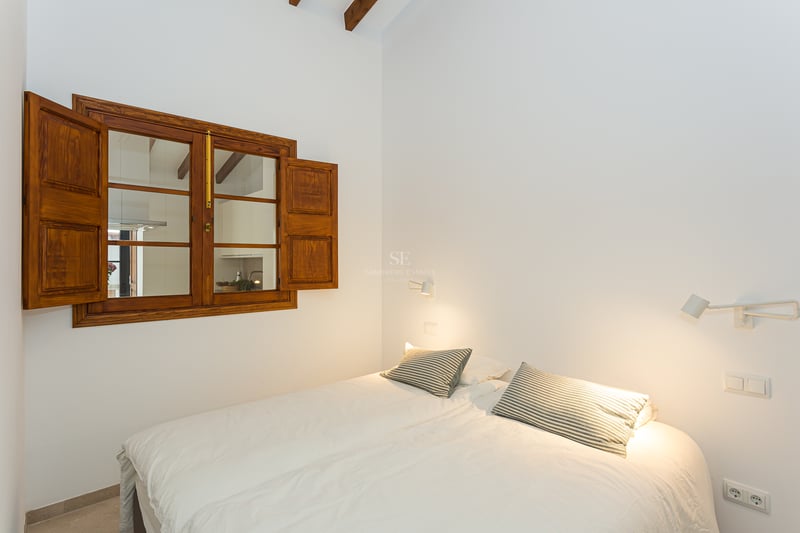 Chambre lumineuse avec literie blanche, oreillers rayés, fenêtre en bois avec volets et poutres apparentes.
