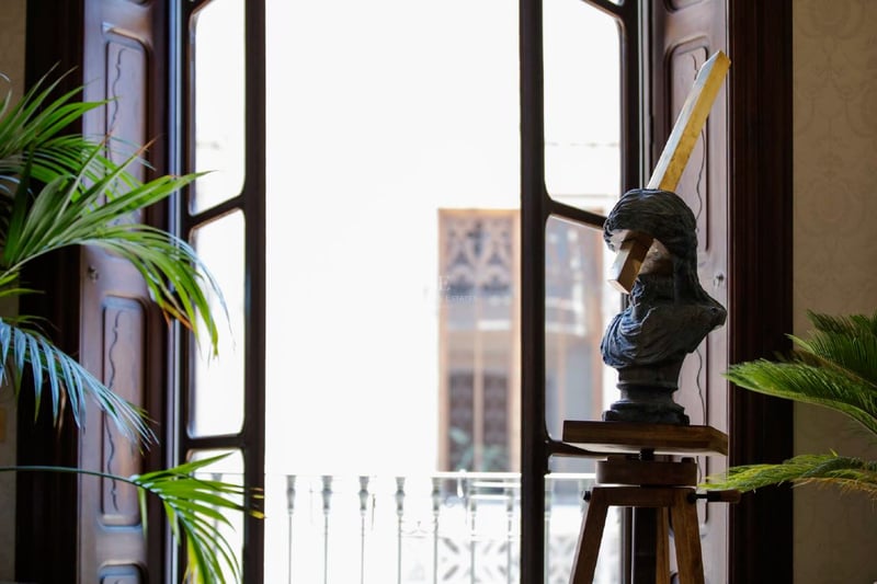 Escultura de un busto sobre un trípode de madera junto a un gran ventanal iluminado con plantas verdes.