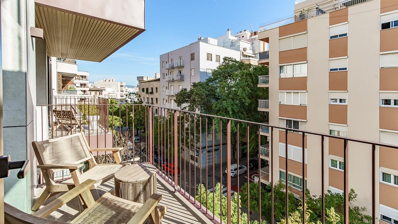 Un balcon ensoleillé avec des chaises et une table en bois surplombant une rue arborée et des immeubles d'appartements.