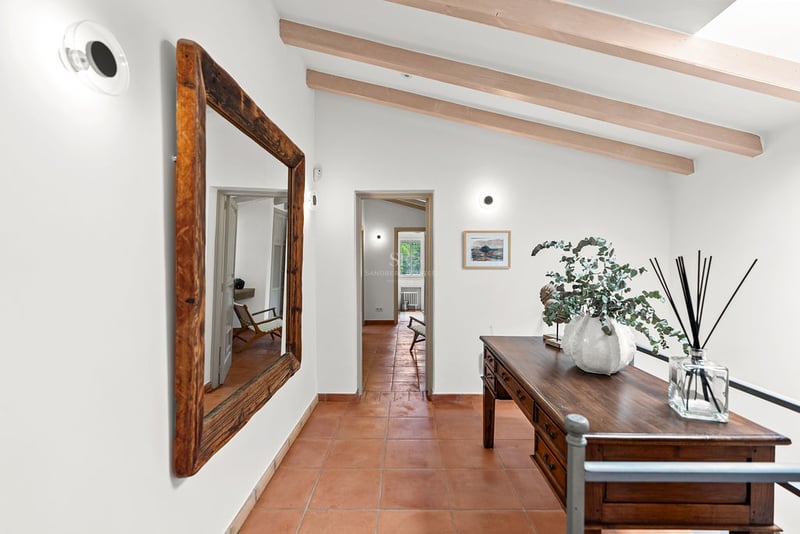 Couloir avec sol en terre cuite, grand miroir en bois, bureau antique et poutres apparentes au plafond.
