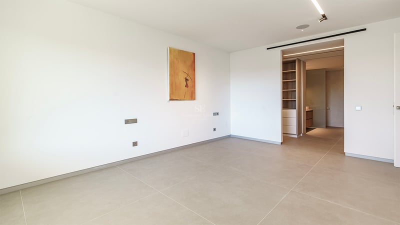 Chambre moderne vide avec grands carreaux beiges, murs blancs, art abstrait et vue sur un dressing en bois.