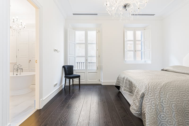 Chambre principale avec parquet foncé, lustre en cristal et vue sur une salle de bain en marbre avec baignoire.