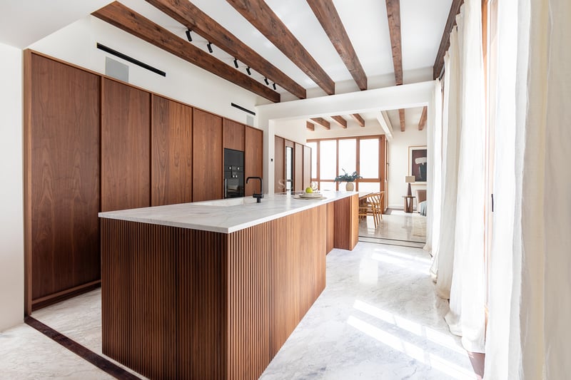 Cocina moderna con isla de madera estriada, encimeras de mármol, vigas de madera vista y luz natural.