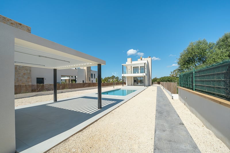 Extérieur d'une villa moderne avec piscine rectangulaire bleue, pergola blanche et cour en gravier sous un ciel clair.