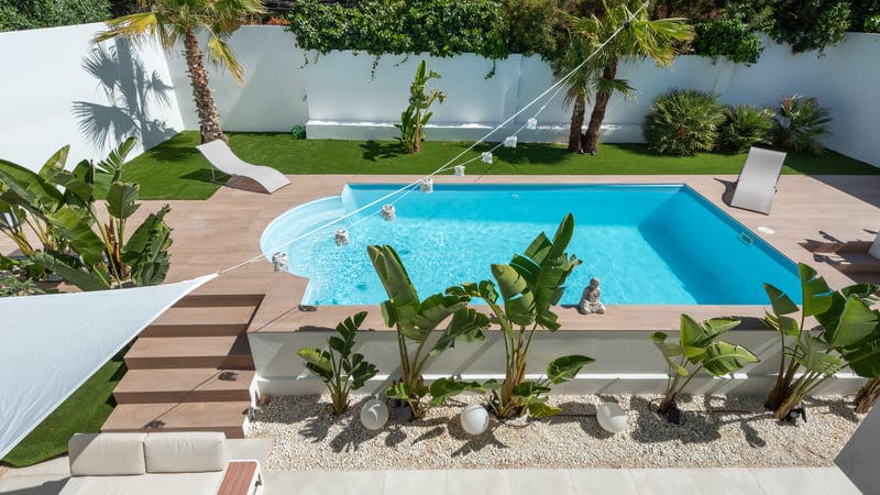 Vue aérienne d'une piscine turquoise entourée de palmiers, de murs blancs et d'une terrasse en grès.
