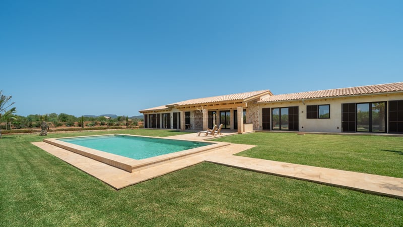 Piscina rectangular rodeada de césped frente a una villa de piedra de una planta bajo un cielo azul despejado.