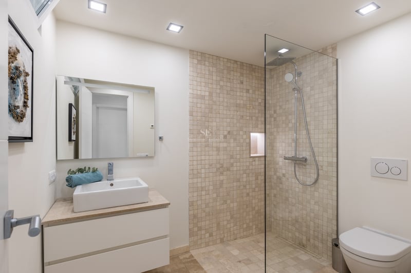 Salle de bains avec douche vitrée, mosaïque en pierre naturelle, meuble vasque blanc et finitions chromées.