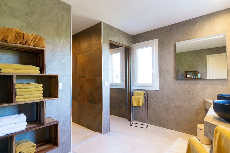 Salle de bain élégante avec murs en pierre grise, étagères en bois avec serviettes jaunes et vasques noires.