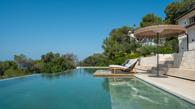 Piscine à débordement avec terrasse en pierre calcaire, bains de soleil et parasol tressé parmi les pins.