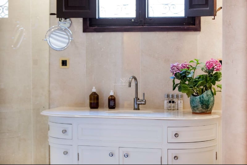 Lavabo blanco con grifo cromado, paredes de mármol beige, espejo de aumento y flores rosas.