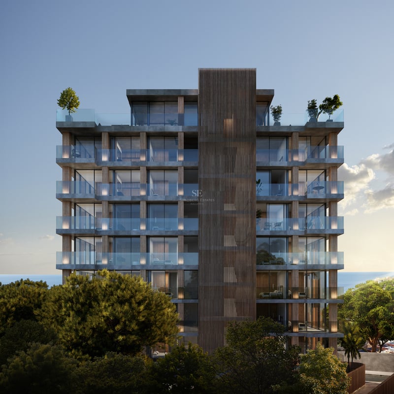 Immeuble moderne de plusieurs étages avec balcons en verre et accents de bois, entouré d'arbres et vue sur la mer.