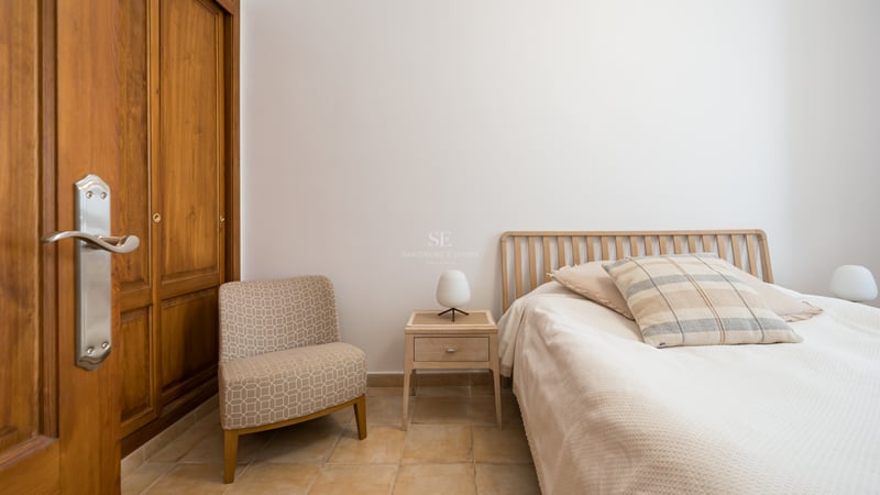 Une chambre lumineuse avec un lit en bois, un fauteuil à motifs, des placards intégrés et un sol en terre cuite.