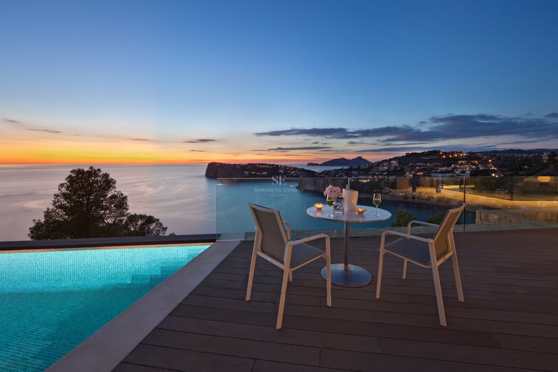 Terrasse de luxe avec piscine à débordement, table pour deux et vue panoramique sur la mer au coucher du soleil.
