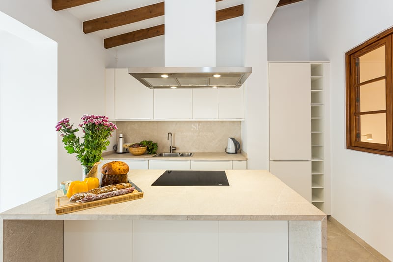 Cuisine lumineuse avec placards blancs, îlot en pierre, poutres en bois apparentes et appareils en acier inoxydable.