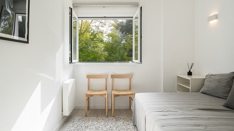 Chambre minimaliste aux murs blancs, chaises en bois et fenêtre ouverte sur une végétation luxuriante.