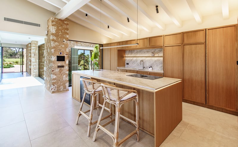 Cocina moderna con isla de madera, encimera de mármol, taburetes de mimbre y techos con vigas blancas.