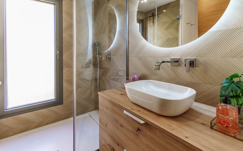 Salle de bain avec vasque à poser sur bois, miroir rond rétroéclairé et paroi de douche en verre.