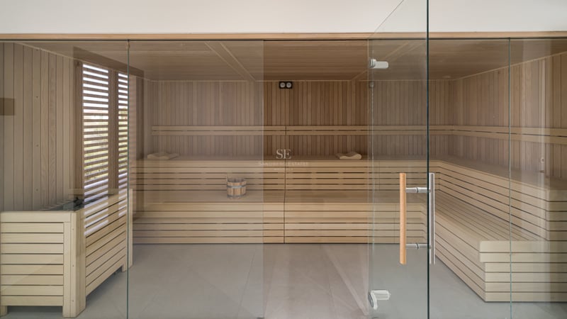 Sauna amplia revestida de madera clara con bancos a varios niveles y una elegante pared frontal de cristal.