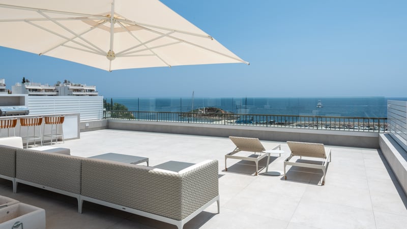 Grande terrasse ensoleillée avec mobilier de salon moderne, garde-corps en verre et vue sur la Méditerranée.