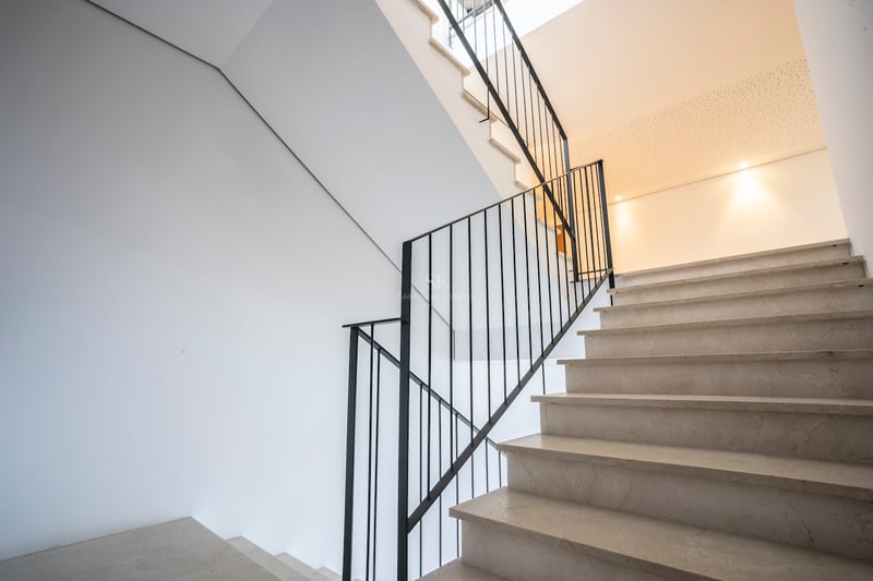 Escalier en marbre crème avec rampe en fer noir contre des murs blancs sous une lumière naturelle douce.