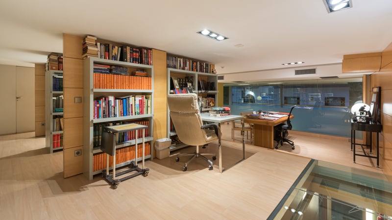 Modern home office with light wood floors, built-in bookshelves, glass desk, and ergonomic leather chair.
