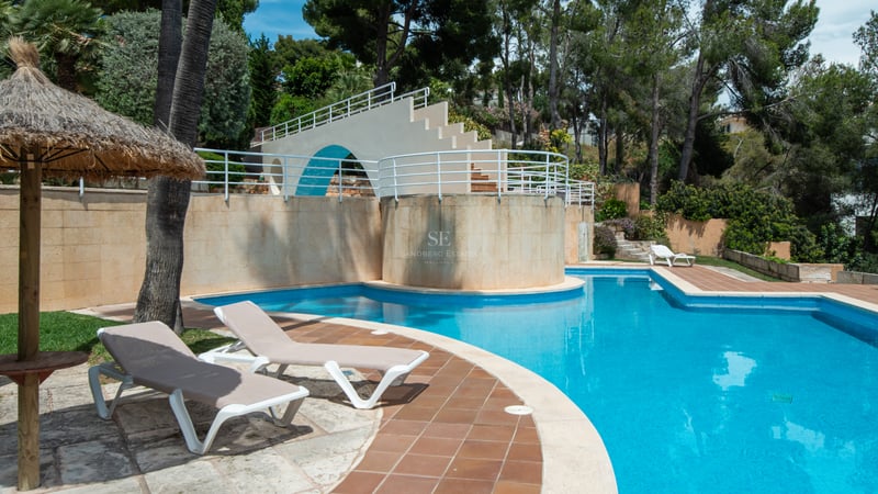 Grande piscine carrelée bleue avec chaises longues, parasol en paille et structure blanche dans un jardin arboré.