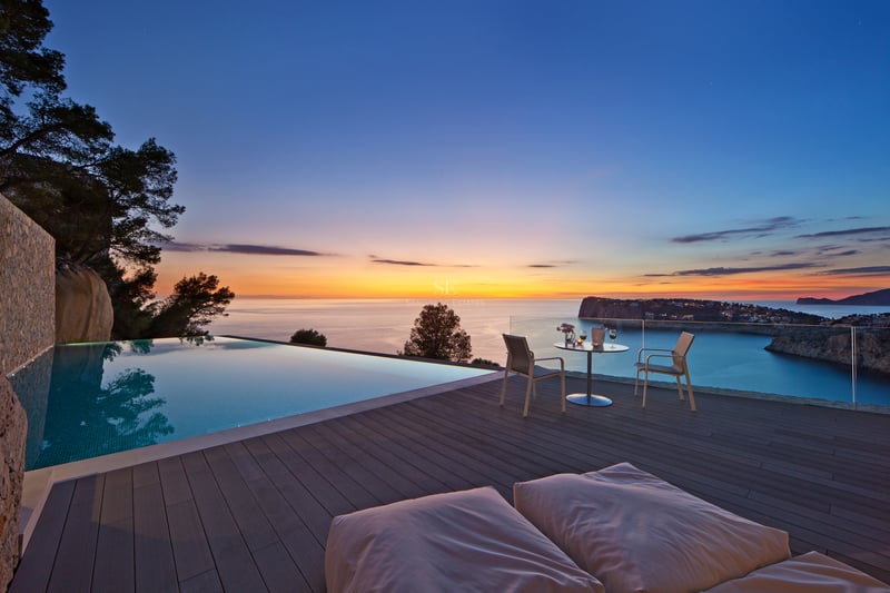 Piscine à débordement sur terrasse en bois avec vue sur la mer au coucher du soleil et mobilier de jardin.
