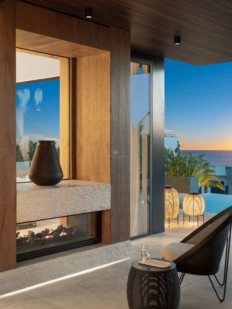 Terrasse de luxe au coucher du soleil avec cheminée vitrée, boiseries en chêne, mobilier design et vue sur la piscine.