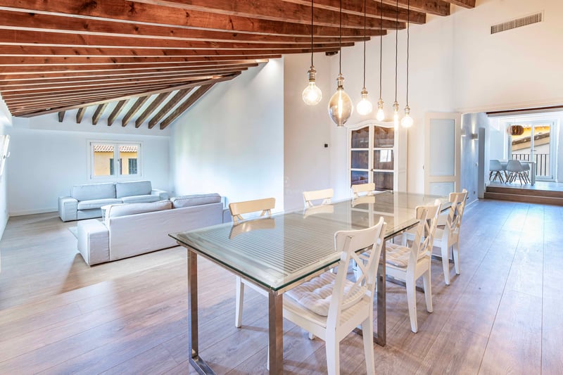 Salle à manger ouverte avec table en verre, poutres en bois au plafond et coin salon en arrière-plan.