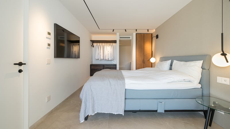 Chambre moderne avec lit bleu, draps blancs, TV murale et dressing ouvert avec éclairage sur rail.