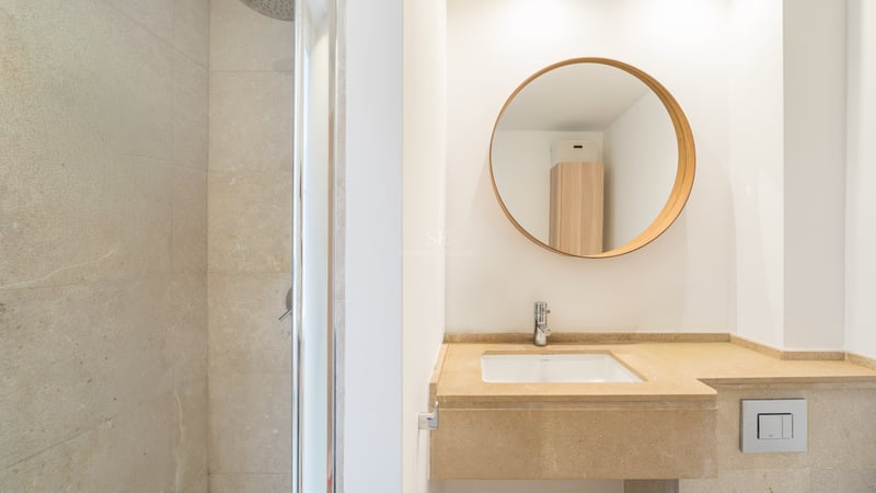 Salle de bain contemporaine avec vasque en pierre beige, miroir rond en bois et douche à l'italienne vitrée.
