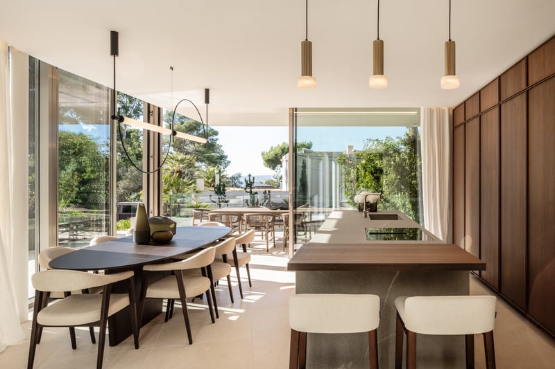 Cuisine de luxe avec îlot en pierre, menuiserie en noyer et salle à manger ouvrant sur une terrasse ensoleillée.