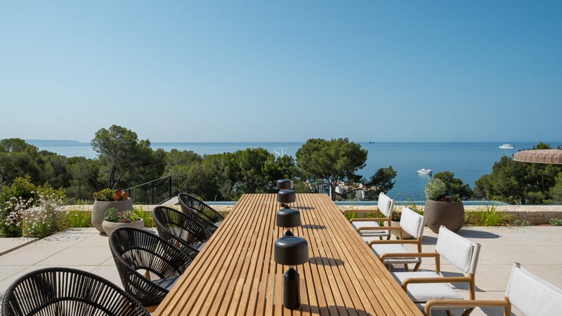 Une table à manger en bois sur une terrasse ensoleillée surplombant la mer Méditerranée et des pins.