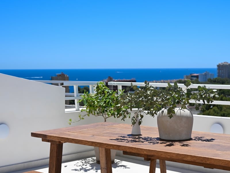 Table en bois rustique sur une terrasse blanche surplombant une mer bleu profond par temps clair.
