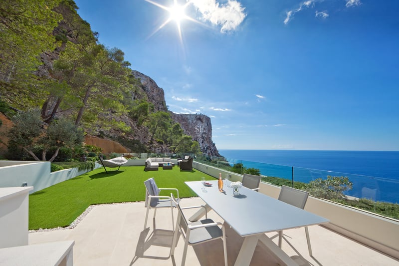 Terrasse moderne avec coin repas, pelouse artificielle et garde-corps en verre face à la mer et aux falaises.