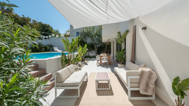 Terrasse moderne blanche avec canapés d'extérieur, coin repas et piscine turquoise sous une voile d'ombrage blanche.
