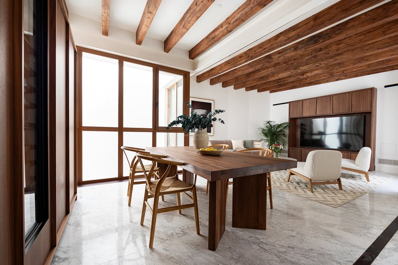 Salón moderno con vigas de madera, suelo de mármol, mesa de comedor de nogal y grandes ventanales.