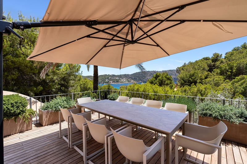 Terrasse extérieure en bois avec table à manger et chaises sous un grand parasol donnant sur la mer.