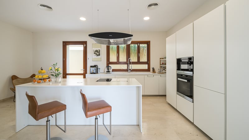 Cuisine blanche moderne avec îlot central, tabourets en cuir, sol en marbre et cadres de fenêtres en bois.