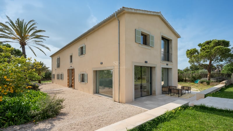 Villa de deux étages couleur sable avec volets vert sauge, chemin en gravier et grandes baies vitrées sous ciel bleu.