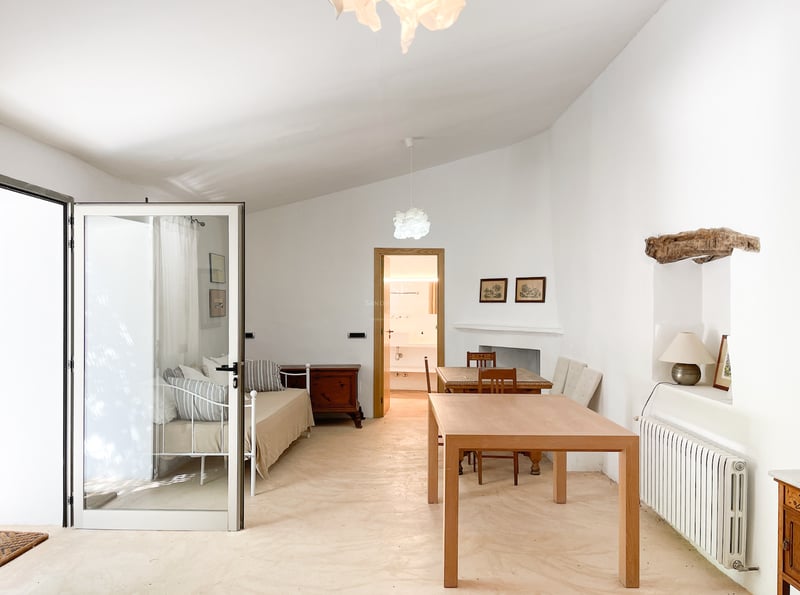 Chambre lumineuse avec lit en métal blanc, table en bois et sol en microciment poli sous un plafond voûté.